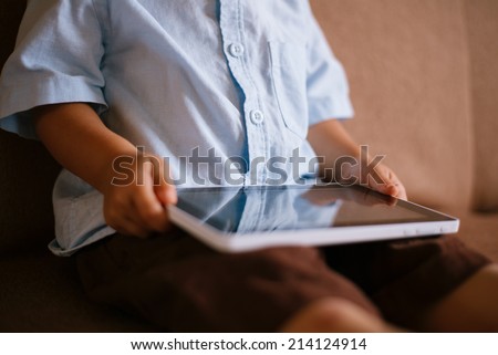Little boy holding a tablet computer on his lap as he sits on the floor leaning against the wall, close up of the tablet