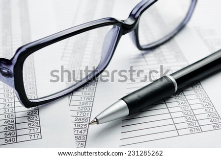 Close up Eyeglasses with Black Frame and Black Ballpoint Pen on Top of Report Papers with Printed Numbers.