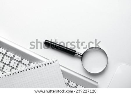Conceptual image depicting conducting an online search for information with a magnifying glass on a blank notebook alongside a wireless computer mouse and keyboard