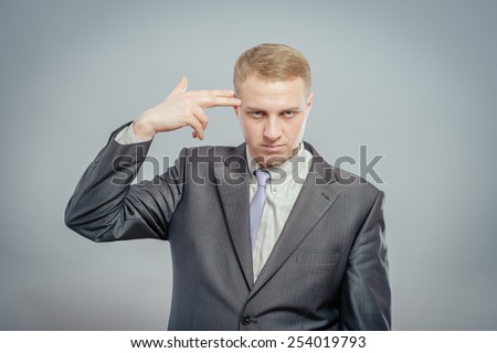 Failed again. Depressed young man in shirt and tie touching his temple with finger gun and looking at camera while standing isolated on gray