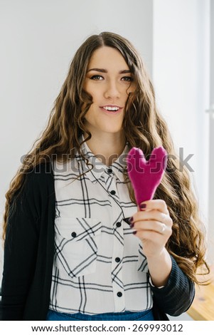 Picture of a small pink heart in hands, lonng hear brunette female holds handmade heart soft toy, woman with Valentine gift against the window, happy girl smiling