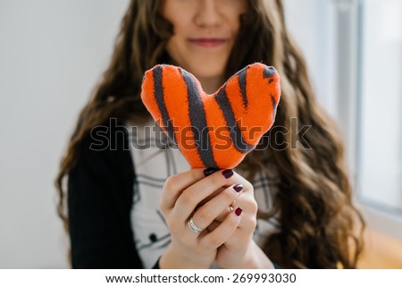 Picture of a small red heart in hands, lonng hear brunette female holds handmade heart soft toy, woman with Valentine gift against the window, happy girl smiling
