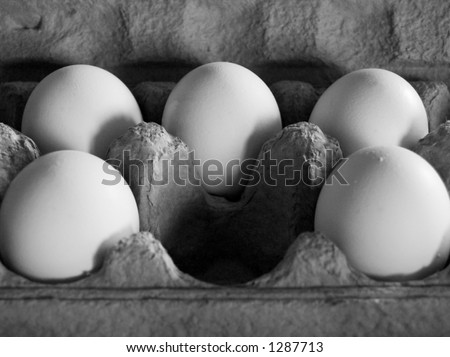 Five eggs in soft, dim light in a grey carton, arranged as to imply that a central egg has been removed. Greyscale image.