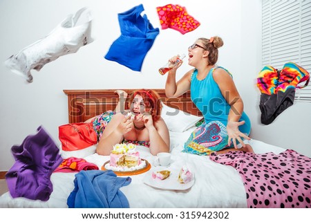 Portrait of beautiful plus size curly red hair young woman eating sweet pastry in bed\
and her sister sitting on bed singing into bottle.