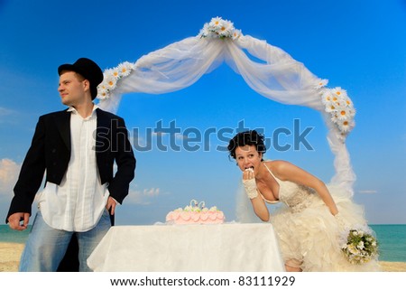 Bride and Groom Under Archway on Beach with wedding cake. Bride trying bit of cake and looking around that nobody see