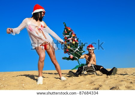 Sexy Santa helper  pulling Santa on a sled with Christmas tree at the beach.  (concept: Tropical winter fun)