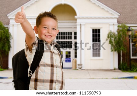 Happy child in front of school wearing back pack and giving thumbs up