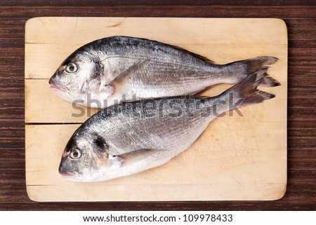 Two fresh sea bream fish on used wooden cutting board on brown background. Culinary seafood eating in natural brown.