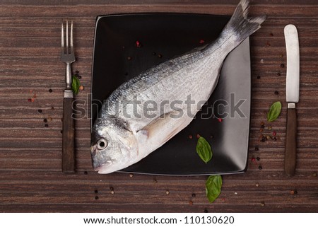 Fresh sea bream on black plate with fresh herbs, colorful peppercorns and wooden cutlery on brown background. Culinary natural seafood background.
