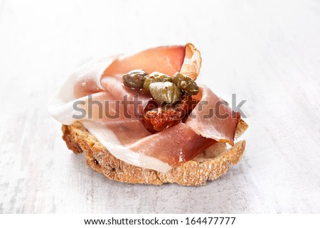Ciabatta piece with prosciutto ham, capers and dry tomatoes on white wooden background. Culinary canape, rustic country style.