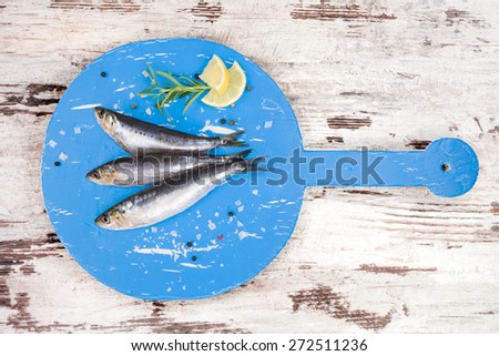 Three fresh anchovy fish on round blue wooden kitchen board on white and brown wooden table, top view. Culinary seafood concept. Delicious healthy eating.