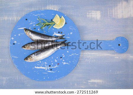Three fresh anchovy fish on round blue wooden kitchen board on blue wooden table, top view. Culinary seafood concept. Delicious healthy eating.