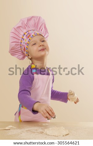 Little girl in chef hat and tablier with classic wooden rolling pin, dough and flour. Gastronomy and culinary cooking.