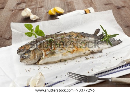 Two grilled trouts with lemon pieces, fresh herbs and garlic on white baking paper on kitchen towel on wooden table.