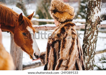 Winter beautiful Girl in Luxury Fur Coat and hat feed the horses