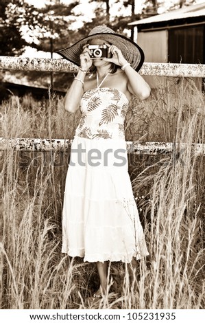 Pretty Woman with Vintage Camera in tall weeds and rusted fence. Photo in old sepia style.