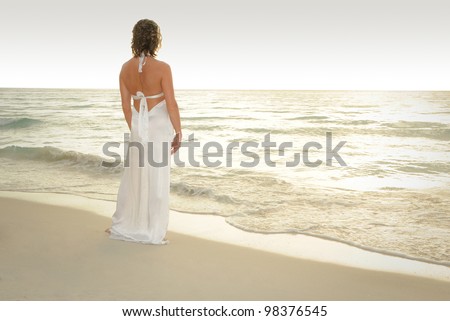 Pretty Woman standing on a white sand beach of a paradise island in a white evening gown looking out at the ocean