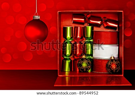 Red Gift Box Filled With Red & Green Party Favors, Decorative Red & White Ribbon And Red, Green & Gold Glitter Christmas Ornaments Over Red Textured Background With LED Lights