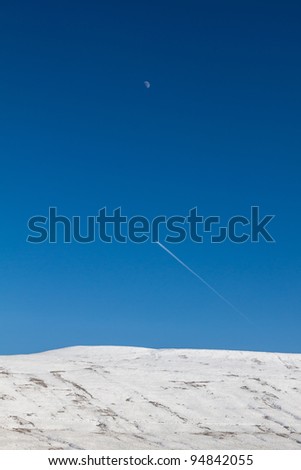 A jet trail in blue sky passes over a snow covered hill with the moon above