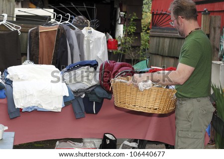 Male holding a basket with linens inside at a yard sale in front of a table with a sleeping bag and clothing items.