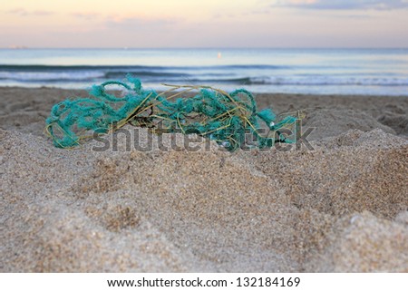 Worn out green blue rope nylon fishing net jumbled up and washed ashore in front of a sandy atlantic coast beach at sunset emphasizing trash\'s endangerment to wildlife.