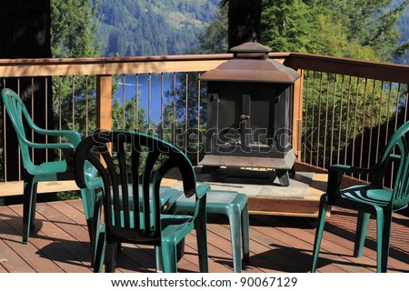 Four plastic chairs on a scenic sunny outdoor deck with a fireplace on a raised platform and a small table all surrounded by a wood and metal railing.