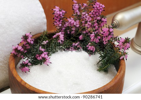 Pink heather flowers in a wood bowl filled with Epsom salts on the top side of a white bath tub near a white cotton towel and silver faucet.