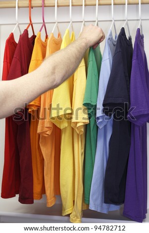 Arm and hand of a caucasian man seen choosing a t-shirt from a variety of shirts in a rainbow of colors in a closet.