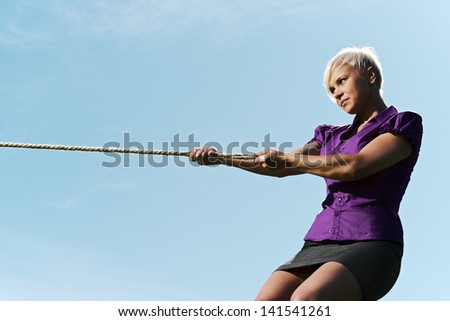 resolute business woman pulling rope against blue sky, symbol of power and determination. Copy space, side view