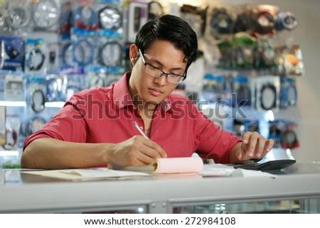Young asian entrepreneur owning a computer shop and checking bills and invoice with calculator, doing budget and reviewing taxes.