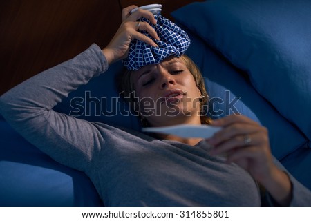 Young hispanic woman in pajamas, laying on bed with fever and ice bag on head, measuring temperature with digital thermometer at night. High angle view