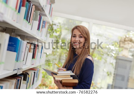 female blonde college student taking book from shelf in library and looking at camera. Horizontal shape, side view, waist up