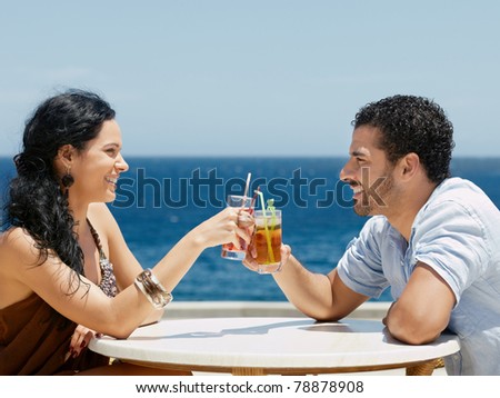 honeymoon: young just married hispanic couple sitting at bar table near the sea and drinking cocktail. Horizontal shape, side view, copy space