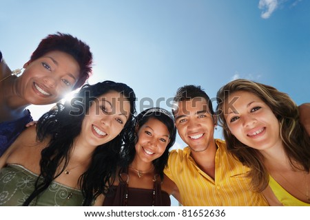 multiethnic group of five male and female friends hugging and looking at camera with sky in background. Low angle view, copy space