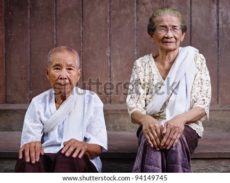 Portrait of two old asian women sitting against brown wall and looking at camera