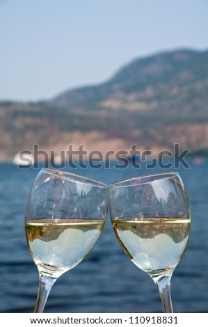 Two wineglasses filled with white wine clinking each other outside on a summer day next to a lake.