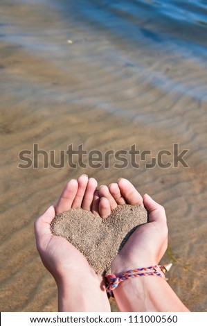 Heart made of sand in a girls hands above crystal clear water at the beach