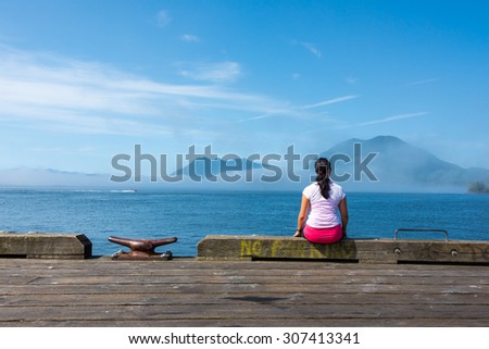 Young woman sitting on edge of dock in Tofino, British Columbia, looking out to the ocean with islands & fog surrounding them.