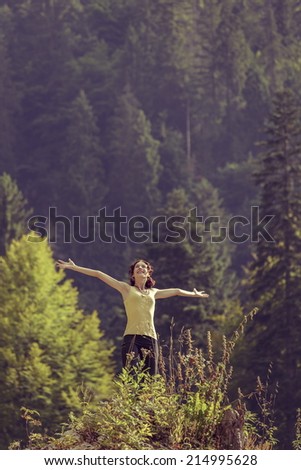 Young delighted woman with arms wide opened, looking up in a gesture of rejoice, freedom and triumph, surrounded by mountain forests.
