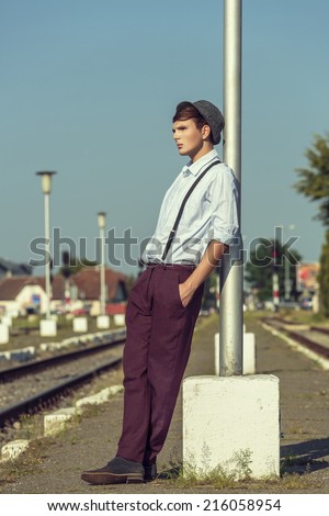 Thoughtful young guy with hand in pocket and rolled up sleeves, wearing hat and trousers with suspenders, leaning his back against a street lamp post while waiting on a railway platform.