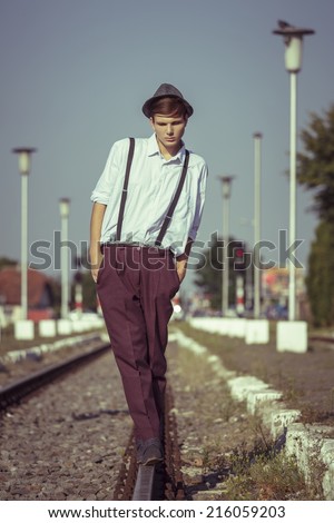 Depressive young guy with hands in pockets and rolled up sleeves, wearing hat and trousers with suspenders, looking down while walking in balance on a rail beside a railway platform.