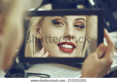 Portrait of a charming smiling woman admiring herself in the mirror. Beauty makeup, skin care and cosmetics treatment. Shallow depth of field and mate color grading.