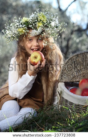 Portrait of adorable kid girl with flower wreath outdoor in the garden. Beautiful girl with curly long hair eating an apple. A child and a basket of apples.