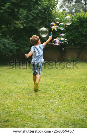 Cute 5 year old boy with brown hair running away from the camera making soap bubbles with garden background