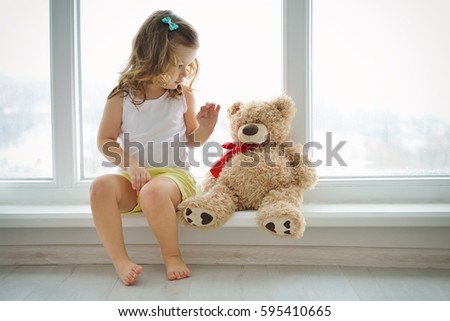 Cute baby at home in white room is sitting near window. The beautiful toddler girl with teddy bear. Baby with curly hair is looking to toy. 商業照片 © 