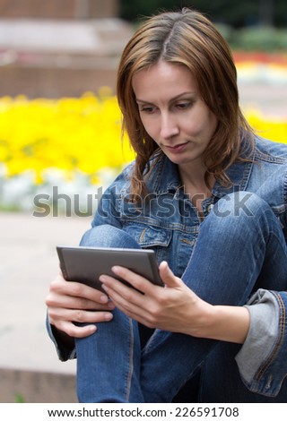 Beautiful attractive cute caucasian young woman reading digital book (ebook) sitting in a park by a summer day with blurred flowers which serve as a background.