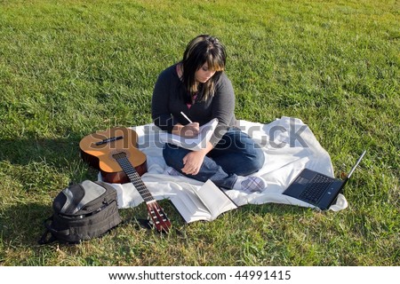 A young female singer or song writer with her guitar and computer outdoors in the grass.