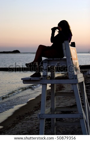 A silhouette of a woman looking with binoculars at the beach while seated in a lifeguard chair.