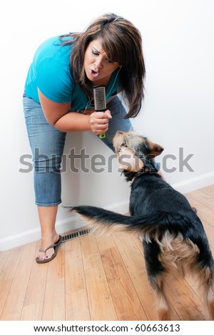 A Hispanic woman singing on a pretend microphone that is actually a hair brush to the dog while listening to music playing through her stereo earbud headphones.