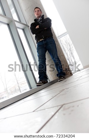A young man standing by some large windows from a low angle perspective.